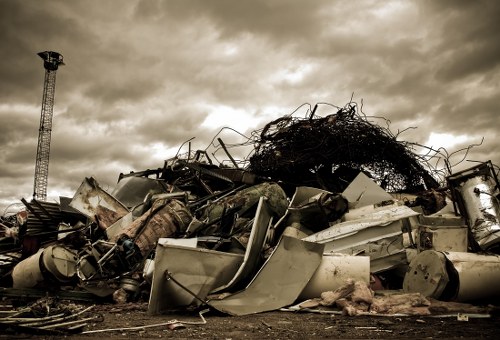 A large pile of discarded, broken appliances and various types of metal debris, including washing machines, refrigerators, and metal frames, is stacked outdoors on a gravel surface with some dirt and smaller fragments mixed in. The metal surfaces show signs of rust and corrosion, with some parts bent or damaged, and the pile is topped by tangled black plastic and wire materials. In the background, a tall metal structure resembling a communications tower or industrial mast is visible against a cloudy, overcast sky with dark, foreboding clouds, creating a dramatic atmosphere. The scene appears to be in an open area, possibly a waste disposal or scrap collection site near Poplar, with a focus on environmental cleanup and rubbish removal services by Commercial Waste Removal Poplar operating within the postcode area.