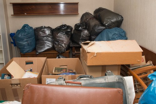 The image shows a cluttered indoor space, possibly a storage room or corner of a building, filled with various waste items ready for removal. Several large black plastic bags are stacked against a beige wall, with some blue bags interspersed among them. In the foreground, an open cardboard box contains miscellaneous small items, and another large cardboard box is partially visible, resting on a wooden surface. A gray plastic garden or construction waste bin lies on its side, partially obscured. Adjacent to the boxes, a small wooden pallet or platform supports some items, including a thick, beige cardboard box with torn edges. The environment appears untidy, with a neutral, utilitarian atmosphere suitable for refuse collection. The setting suggests a site where Commercial Waste Removal Poplar provides rubbish clearance, taken in a residential or commercial context within the London Borough of Tower Hamlets, accessible via postcode E14 or nearby areas.